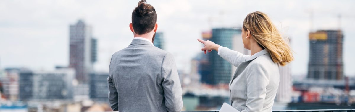 Rear view of two cheerful young business people with laptop standing on a terrace outside office, talking.